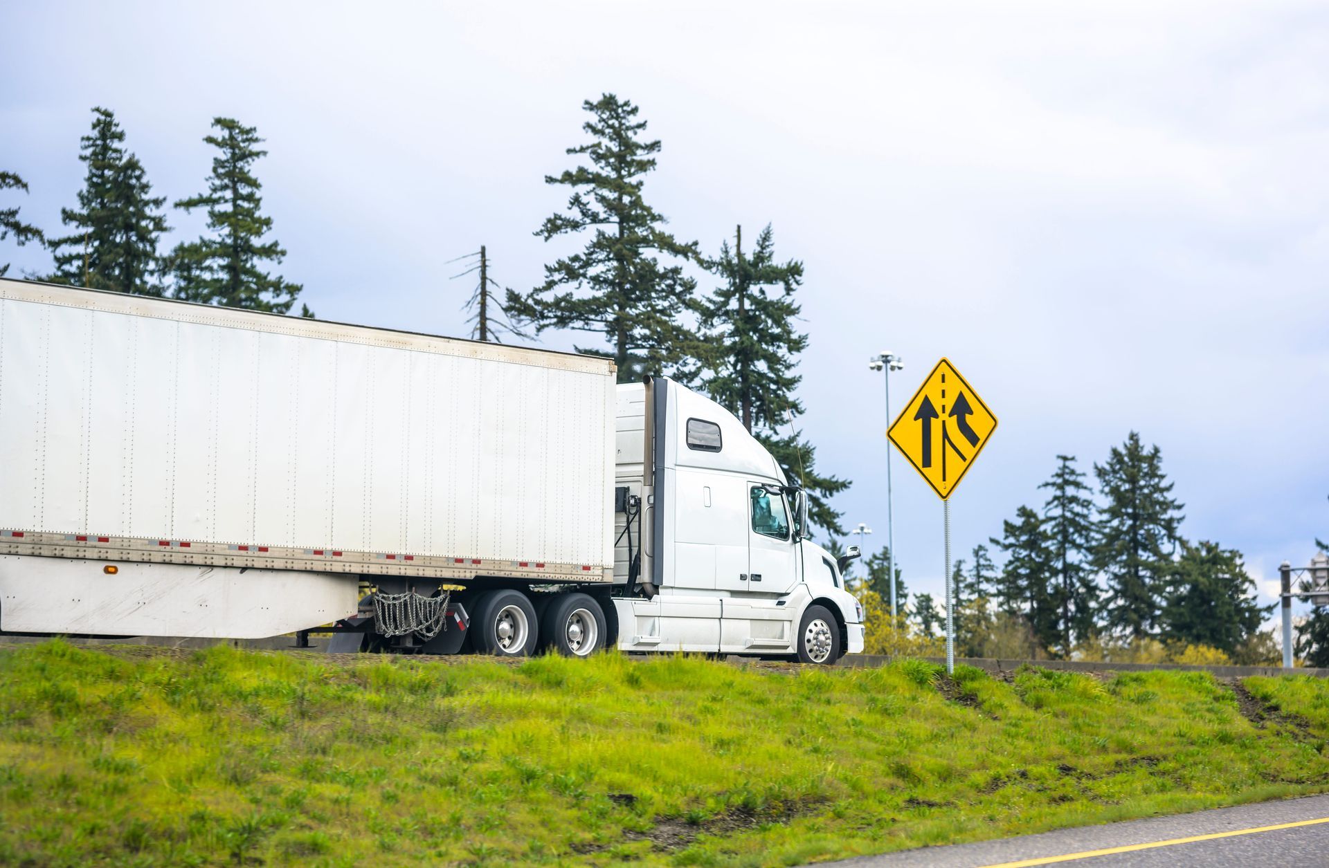 White semi-truck on grassy roadside near a yellow merge sign and pine trees