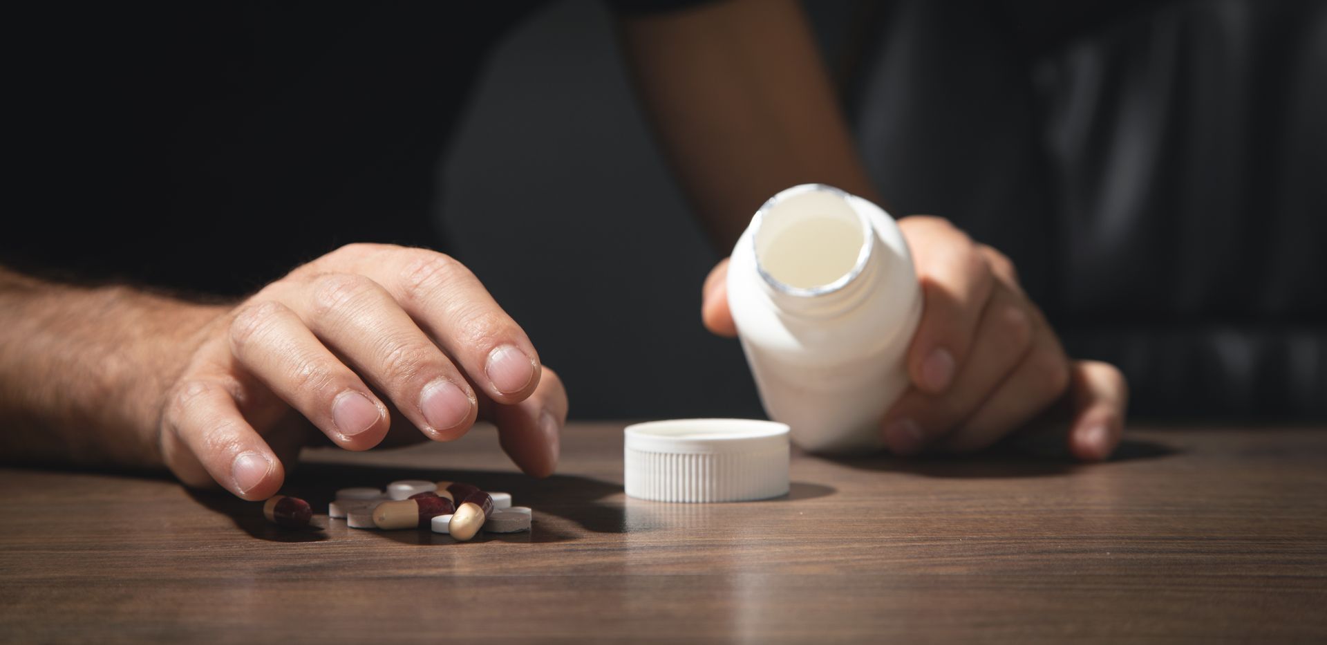 Person with pills and bottle on table; potential overdose, pills scattered, hand reaching.