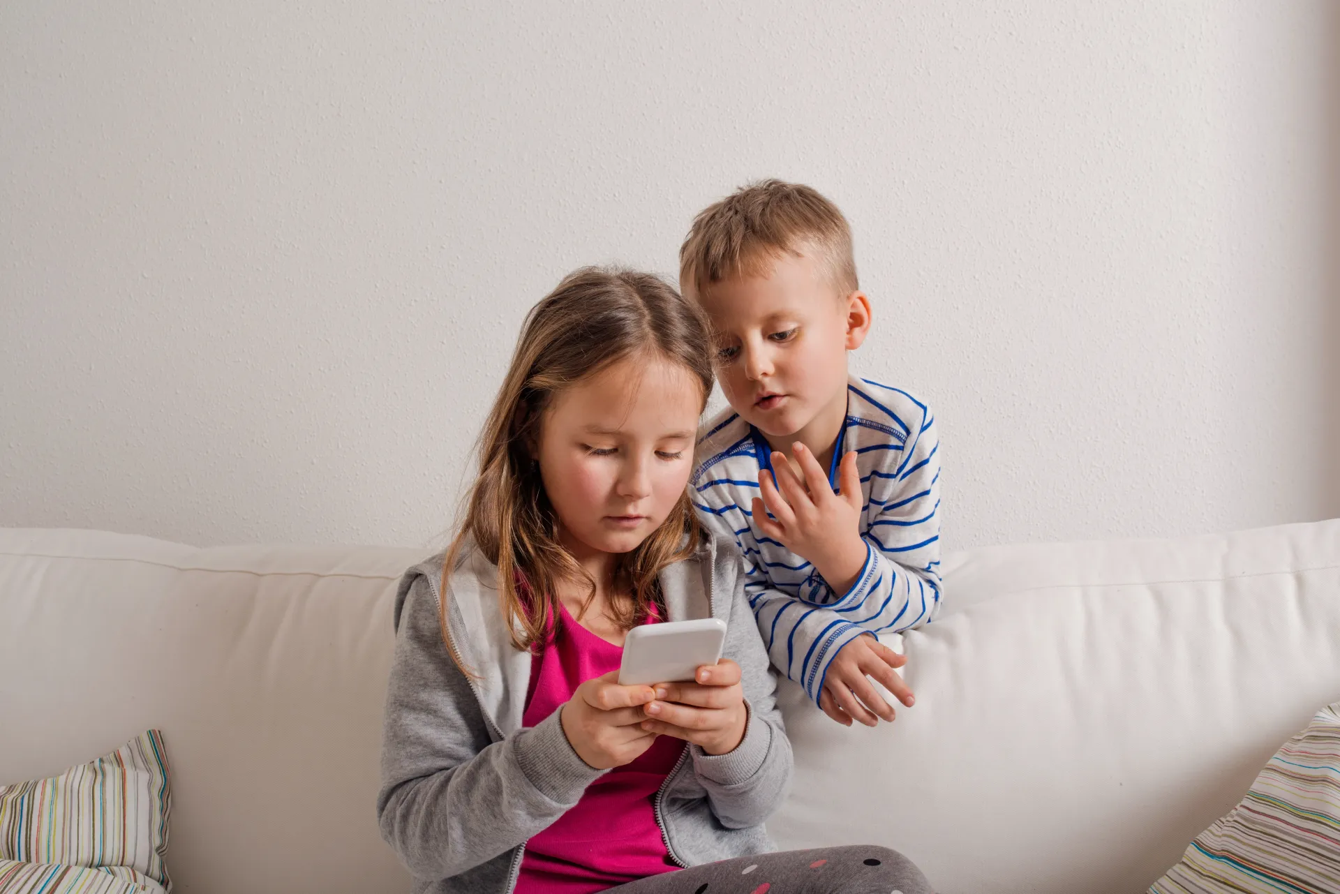 Girl on sofa looks at phone. Boy peers over her shoulder, surprised. White background.