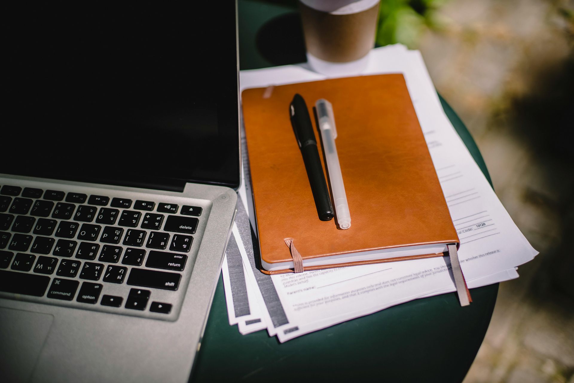 Laptop, notebook with pens, papers, and coffee cup on a green table outdoors.
