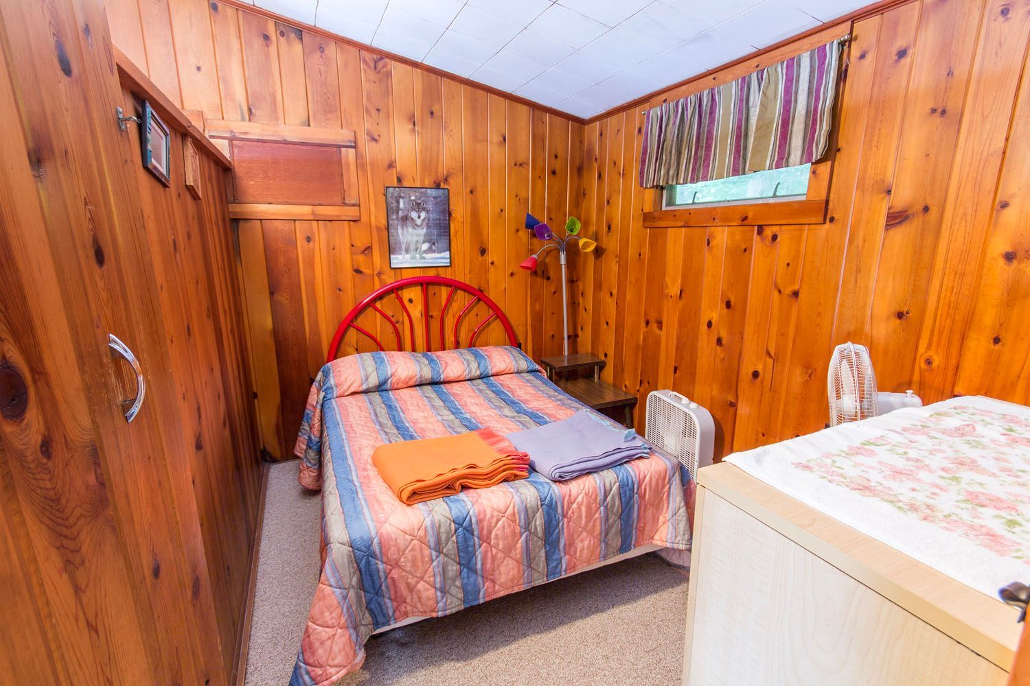 A bedroom with a bed , dresser , fan and wooden walls