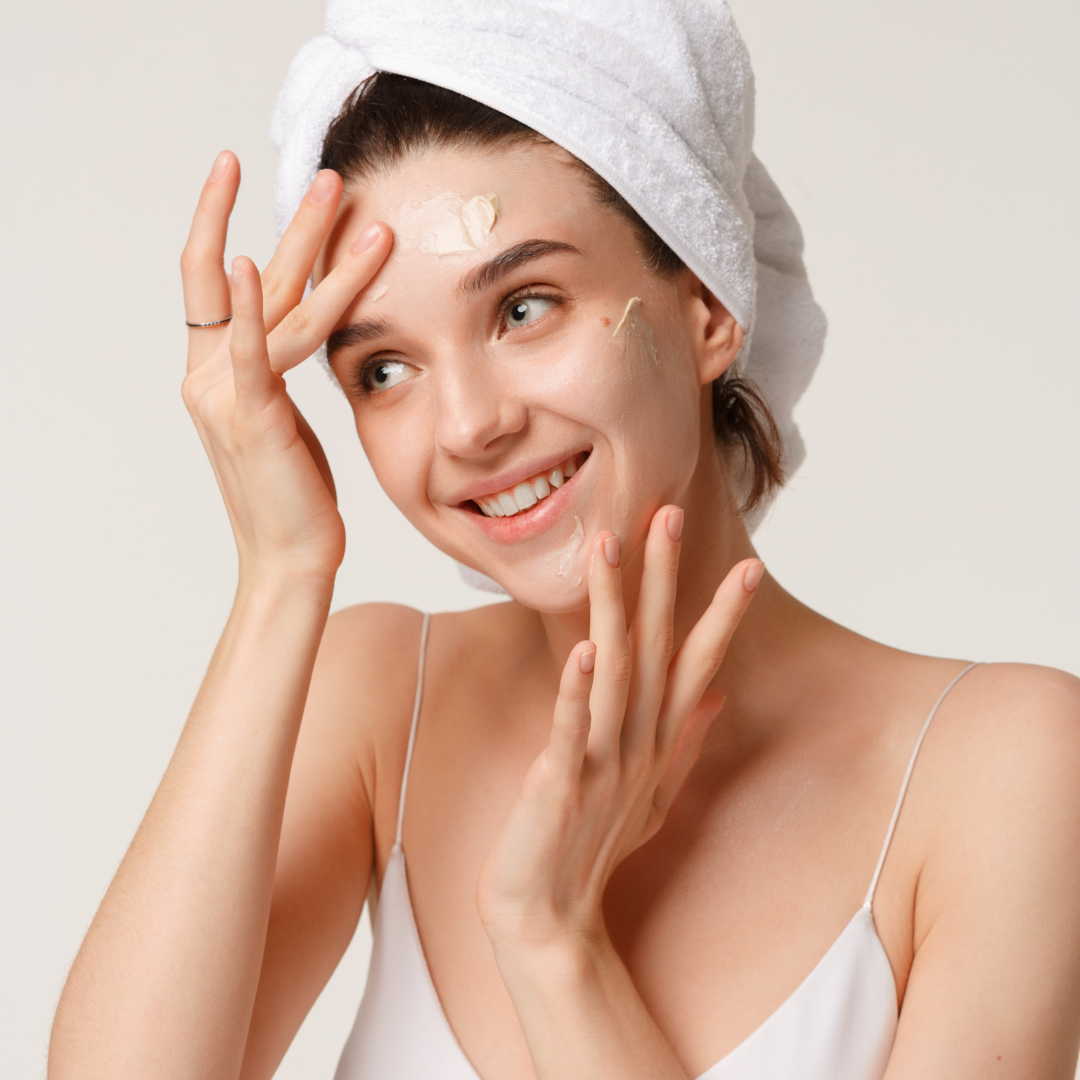 A woman with a towel wrapped around her head is applying lotion to her face.