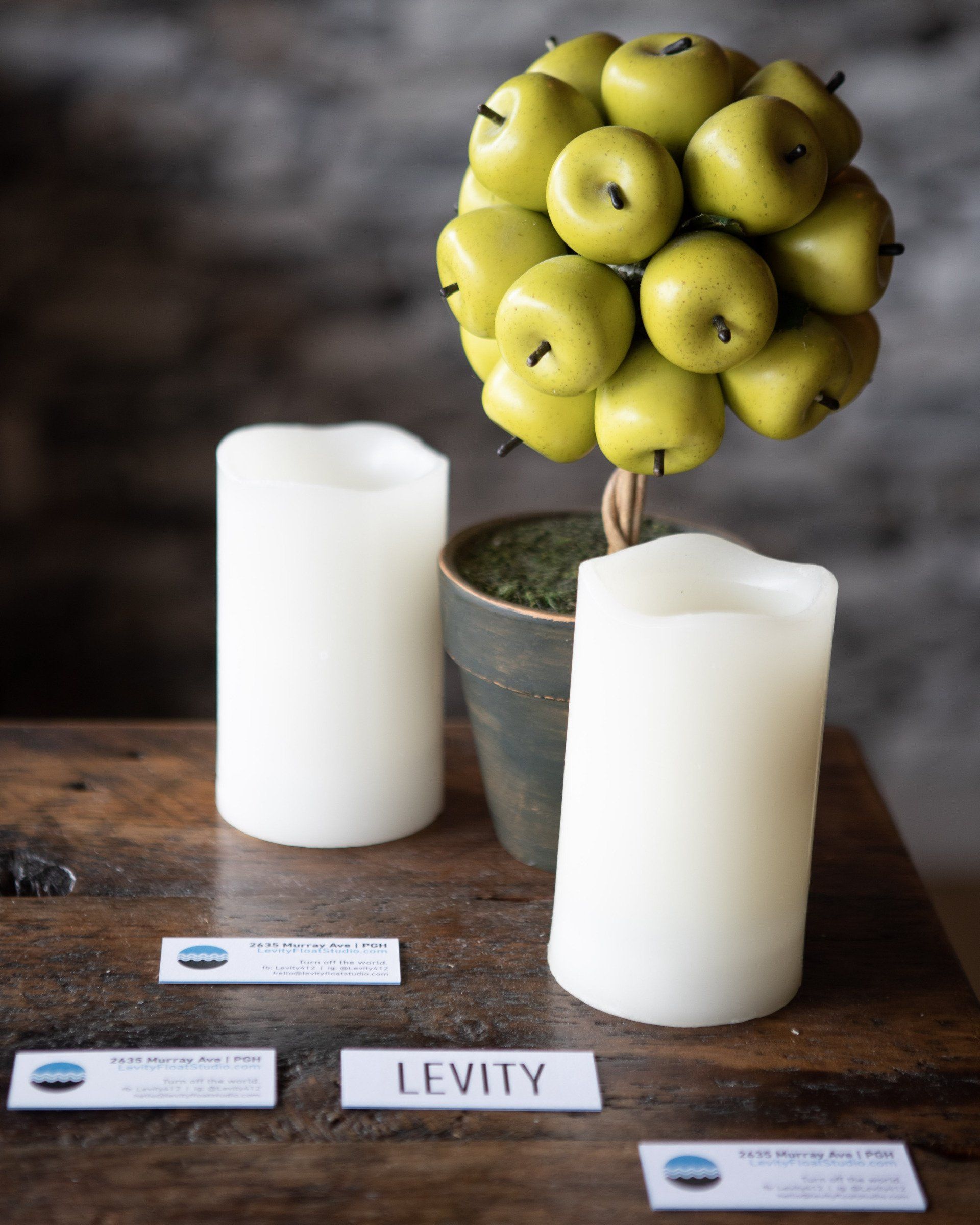 Two candles are sitting on a table next to a topiary of apples and a sign that says levity