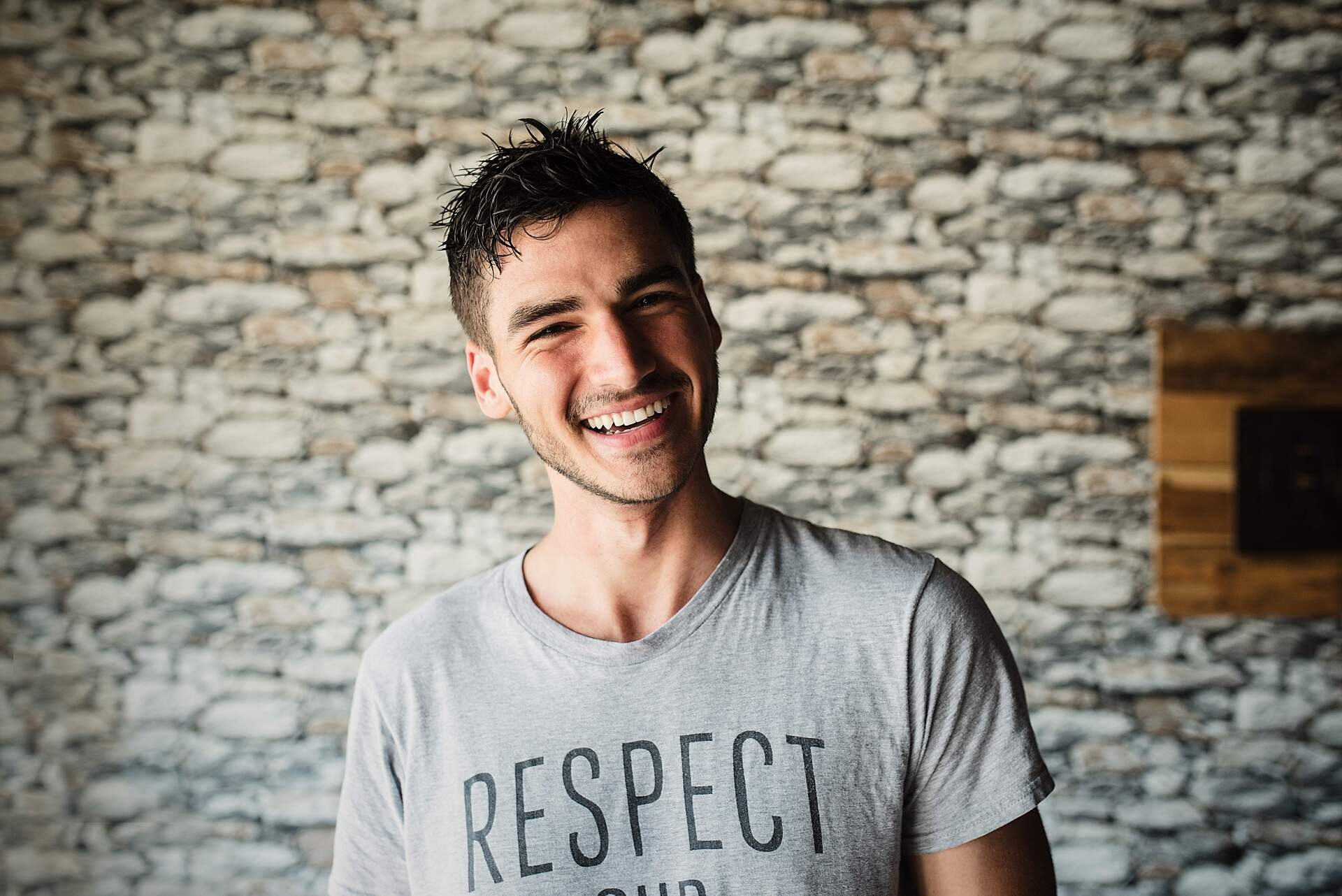 A man wearing a respect t-shirt is smiling in front of a stone wall.