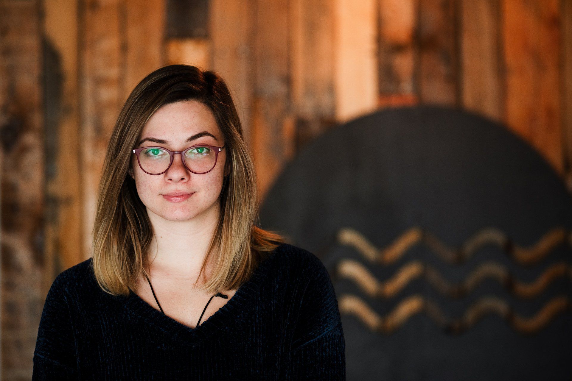 A woman wearing glasses and a black sweater is standing in front of a wooden wall.