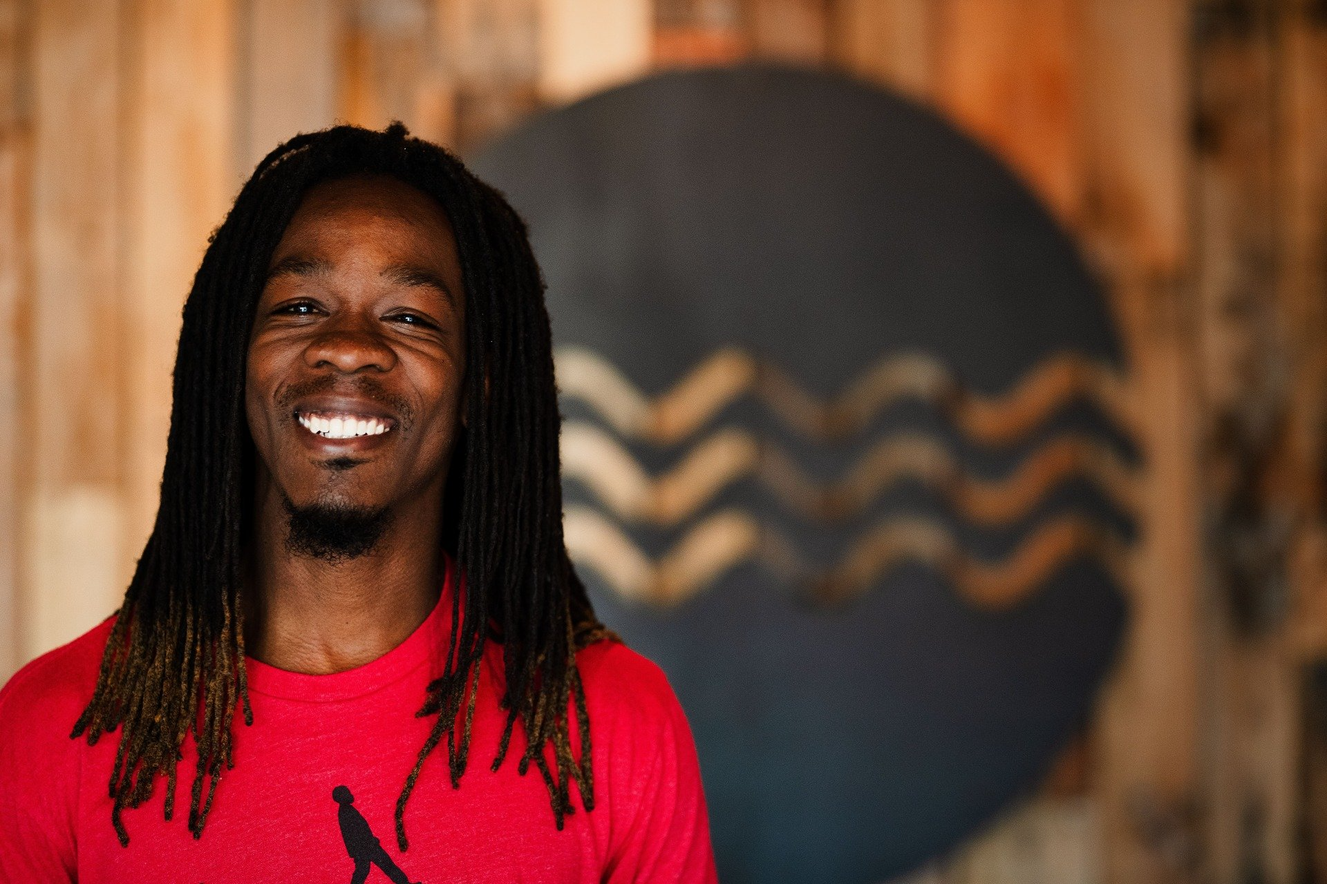 A man with dreadlocks is smiling in front of a wooden wall.