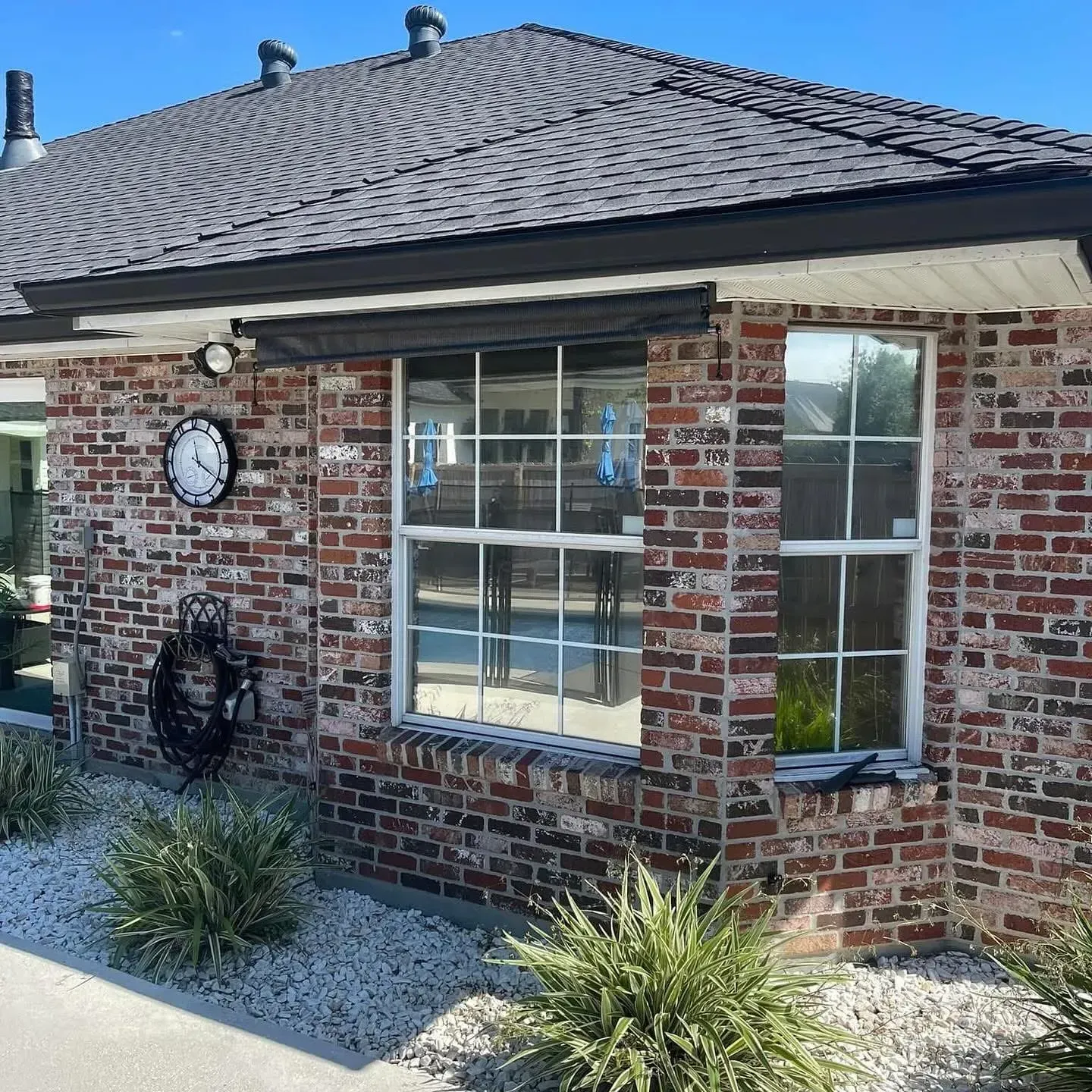Brick house exterior with a black roof and white-framed windows, shrubbery, and a wall clock.
