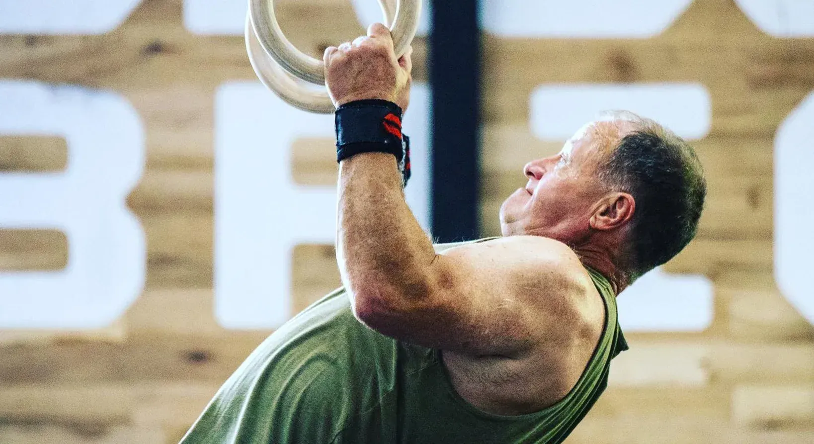 Man doing ring pull-ups at Brigade Athletic Club in Chattanooga, wearing wrist supports, in a gym with wooden paneling.