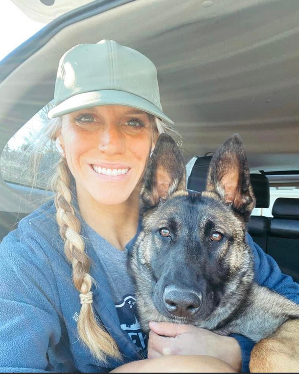 Woman in a cap smiles next to a dog in a car. The dog is brown and black, and has its head close to the woman's face.