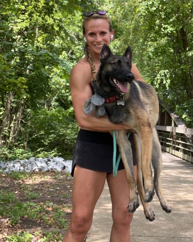 Woman holding a tan and black dog on a trail, smiling. She wears black shorts and sunglasses. Trees in the background.