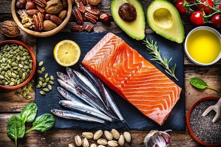 Healthy foods, including salmon, avocado, nuts, seeds, and oil, arranged on a wooden table.