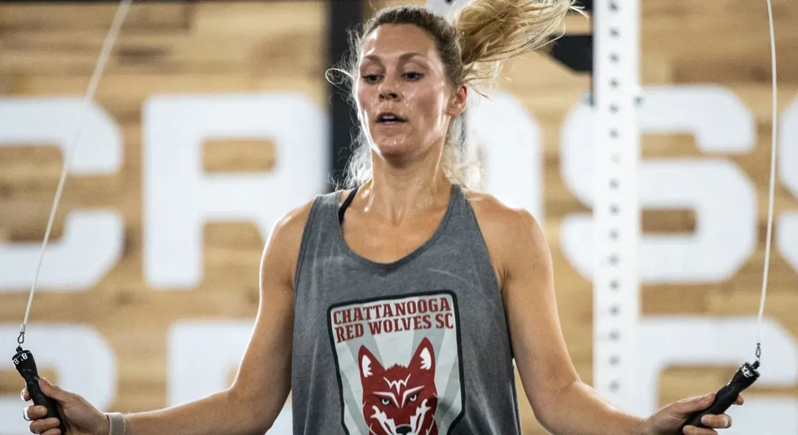 Woman at Brigade Athletic Club in Chattanooga jumping rope, wearing a gray tank top, sweat visible, blurred background.