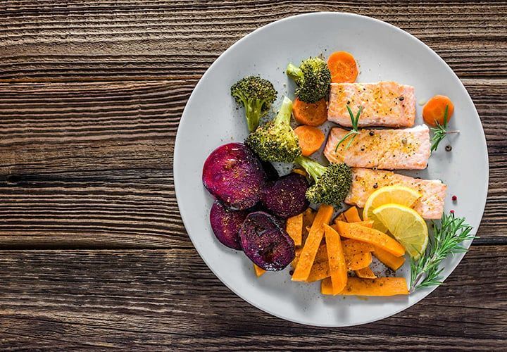 Plate of cooked salmon, vegetables (broccoli, carrots, sweet potatoes, beets), and lemon on a wood table.