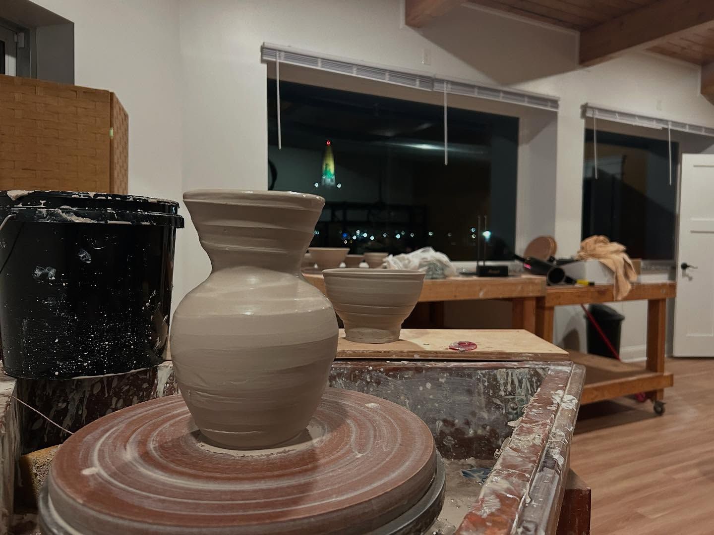 A pottery wheel with a clay vase taking shape; workshop setting, other works in the background, nighttime through windows.