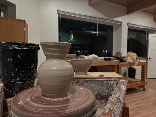 A pottery wheel with a clay vase taking shape; workshop setting, other works in the background, nighttime through windows.