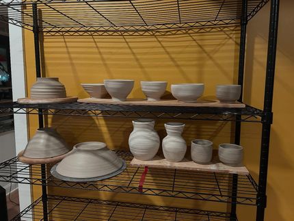 Pottery displayed on shelves, including bowls, vases, and cylindrical forms; the background is a yellow wall.