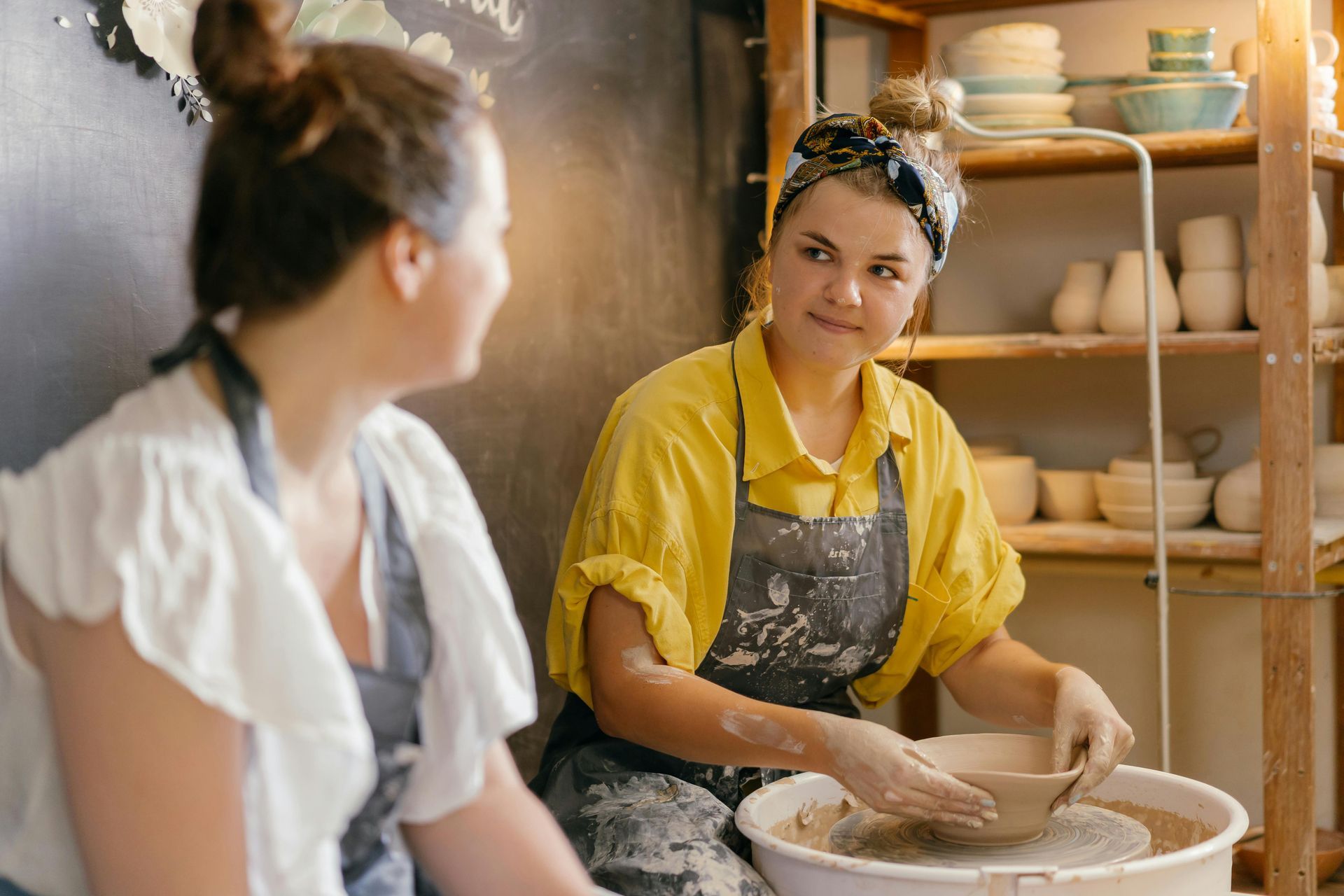 Woman in yellow shirt shapes pottery on a wheel while another woman watches in a workshop.