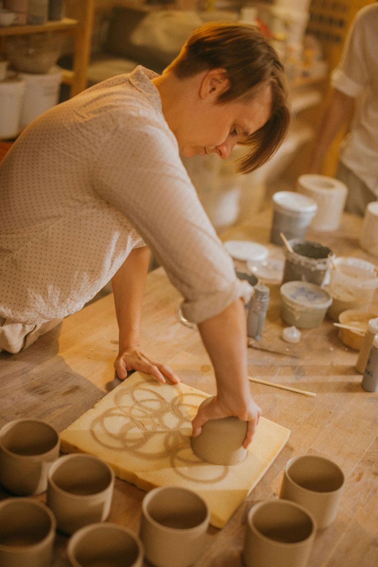 Man teaching woman pottery on a wheel; both are smiling and covered in clay.