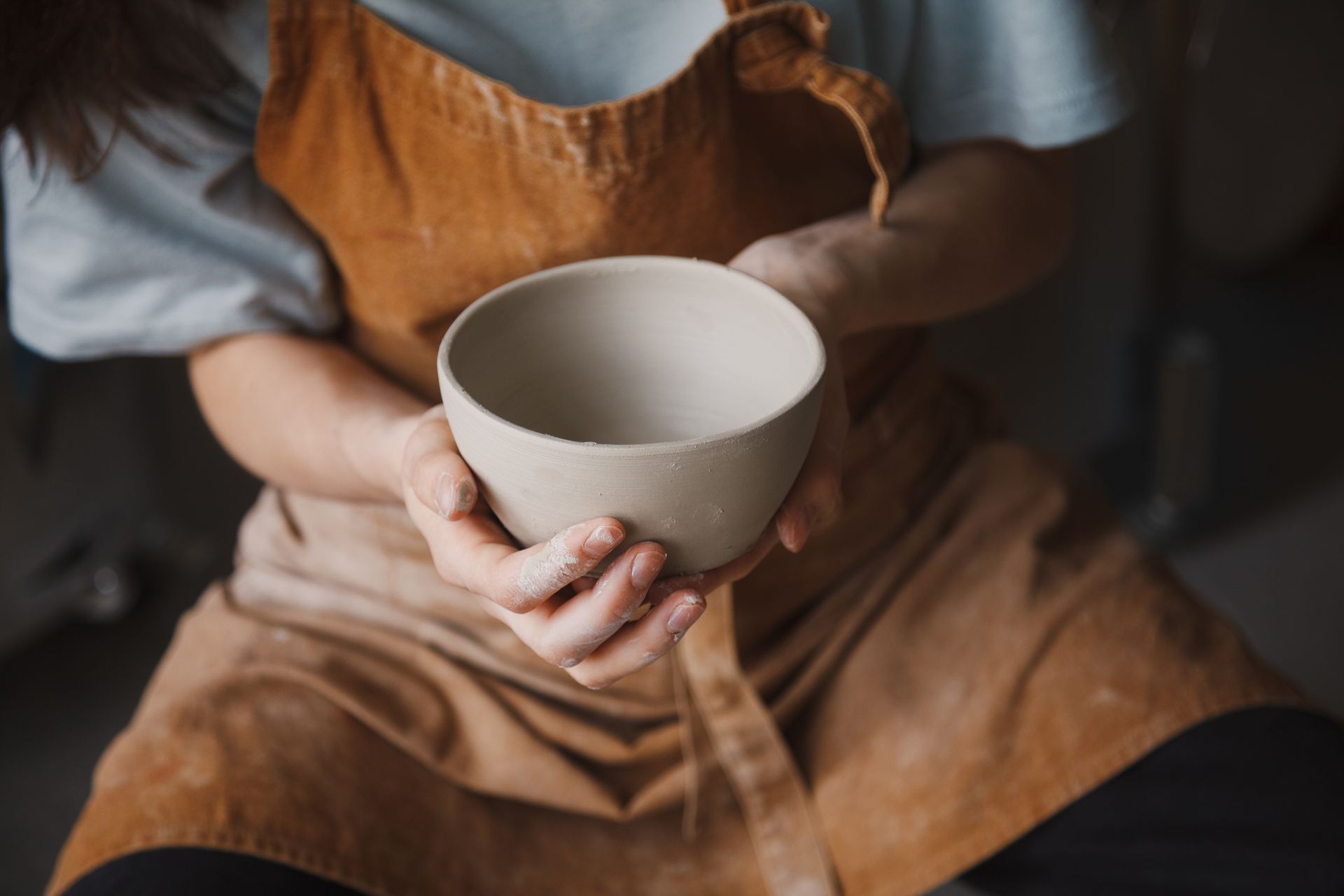 Person holding a light-colored, unfinished clay bowl; hands are covered in clay, wearing an apron.