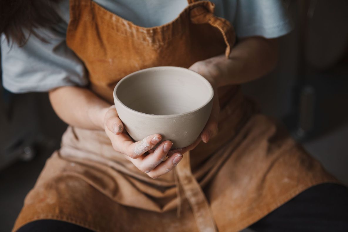 Person holding a light-colored, unfinished clay bowl; hands are covered in clay, wearing an apron.