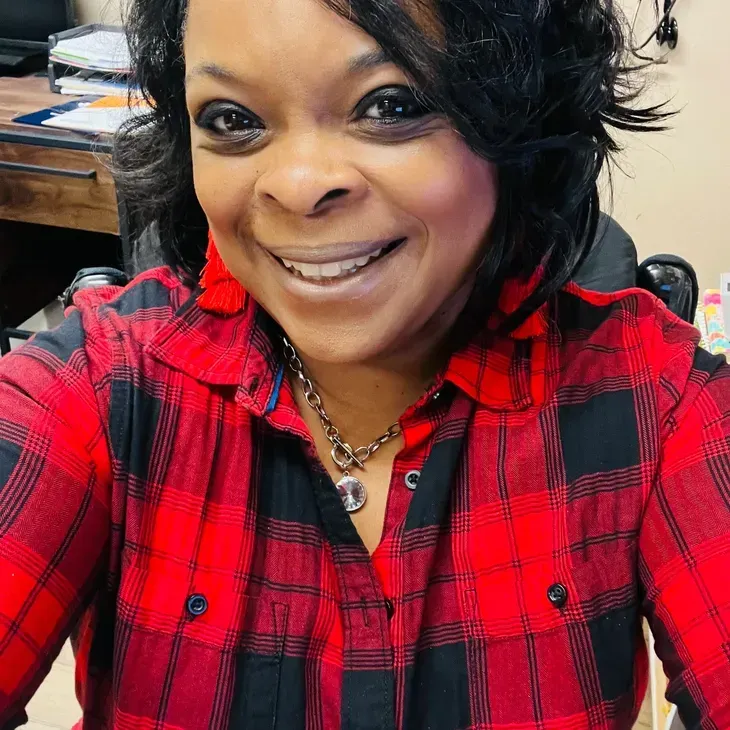 Woman with short black hair smiles wearing red and black plaid shirt, red earrings, indoors.
