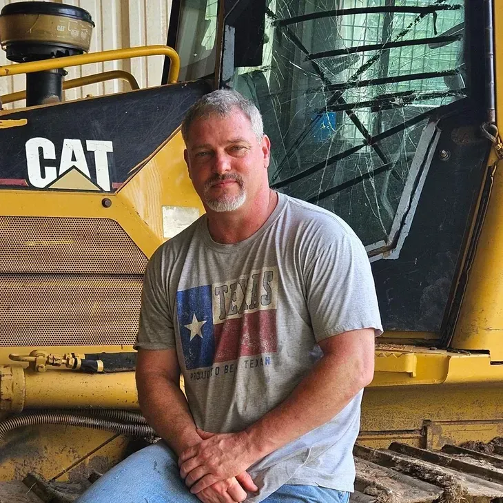 Man in gray t-shirt sitting on a yellow CAT bulldozer, with Texas flag on shirt.