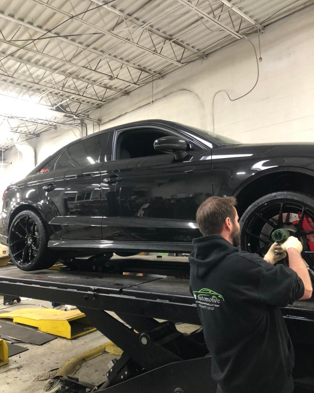 Man working on a black car lifted on a hoist in a garage, holding a tool near the wheel. | SB Automotive