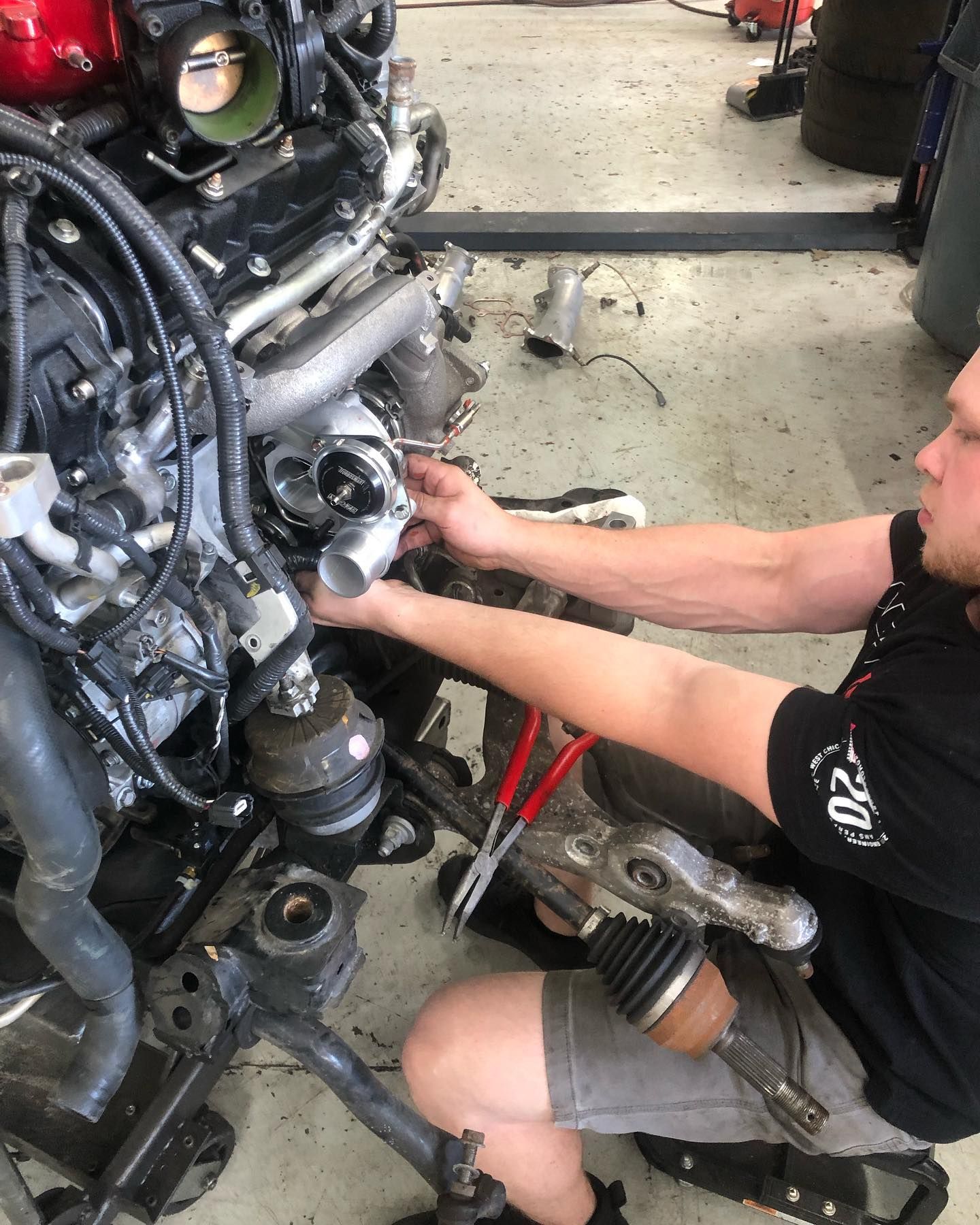 Mechanic working on a car engine. He's holding pliers, surrounded by parts, in a garage setting. | SB Automotive
