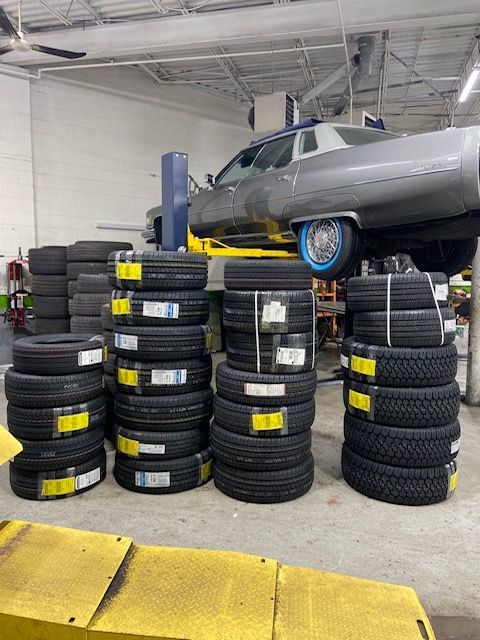 Stacks of new tires in a shop with a car raised on a lift, waiting for service. | SB Automotive