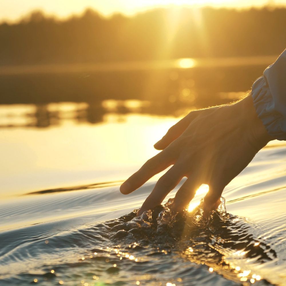 A person 's hand is touching a body of water at sunset