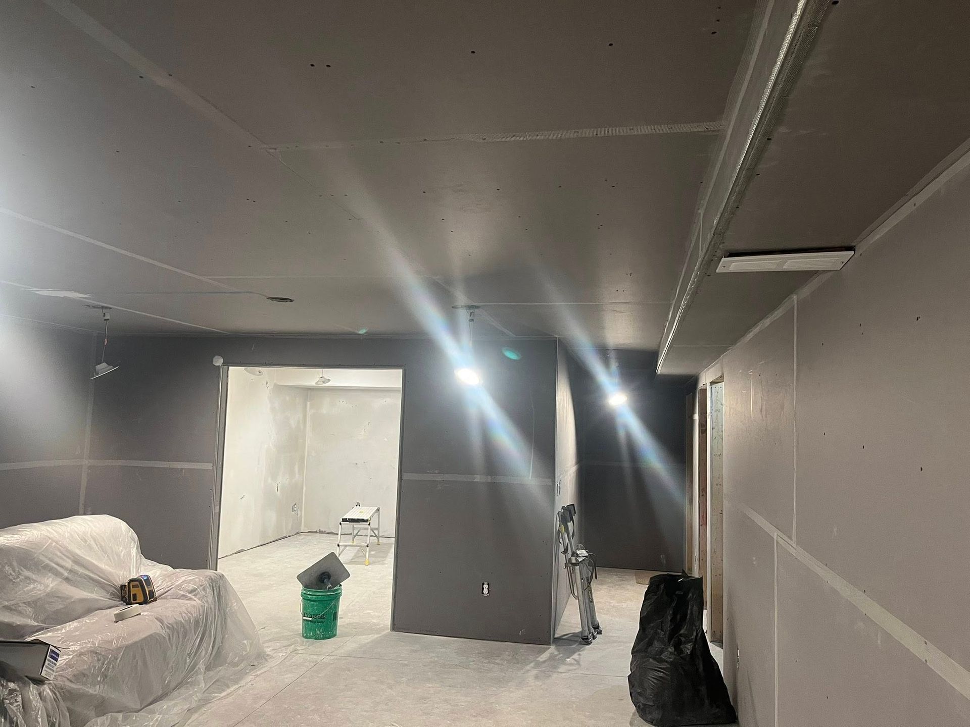 A basement interior under construction, featuring gray drywall on the walls and ceiling, a plastic-covered sofa, and tools.