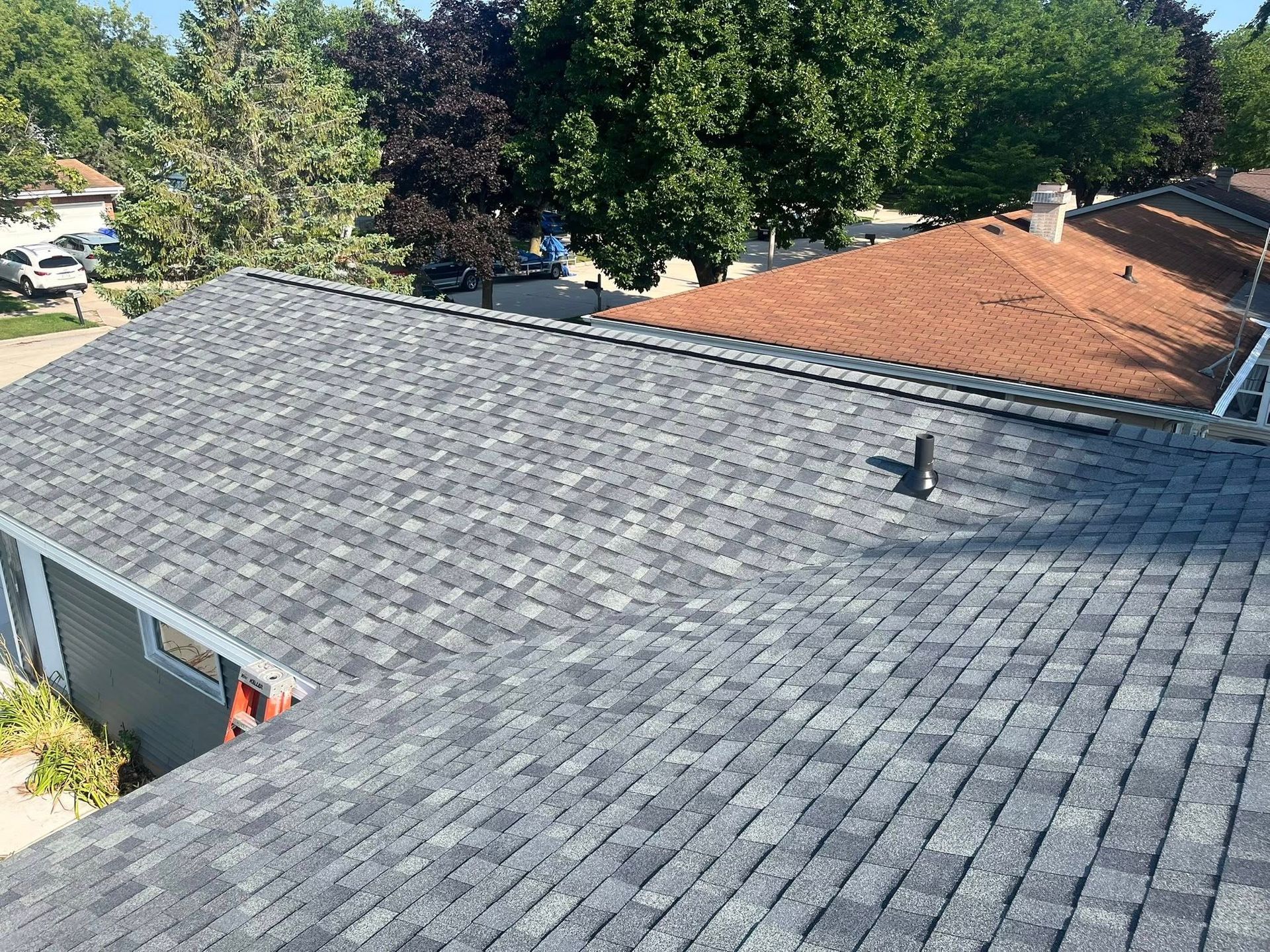 A high-angle view of two adjacent residential roofs with gray and brown shingles under a bright, sunny sky.