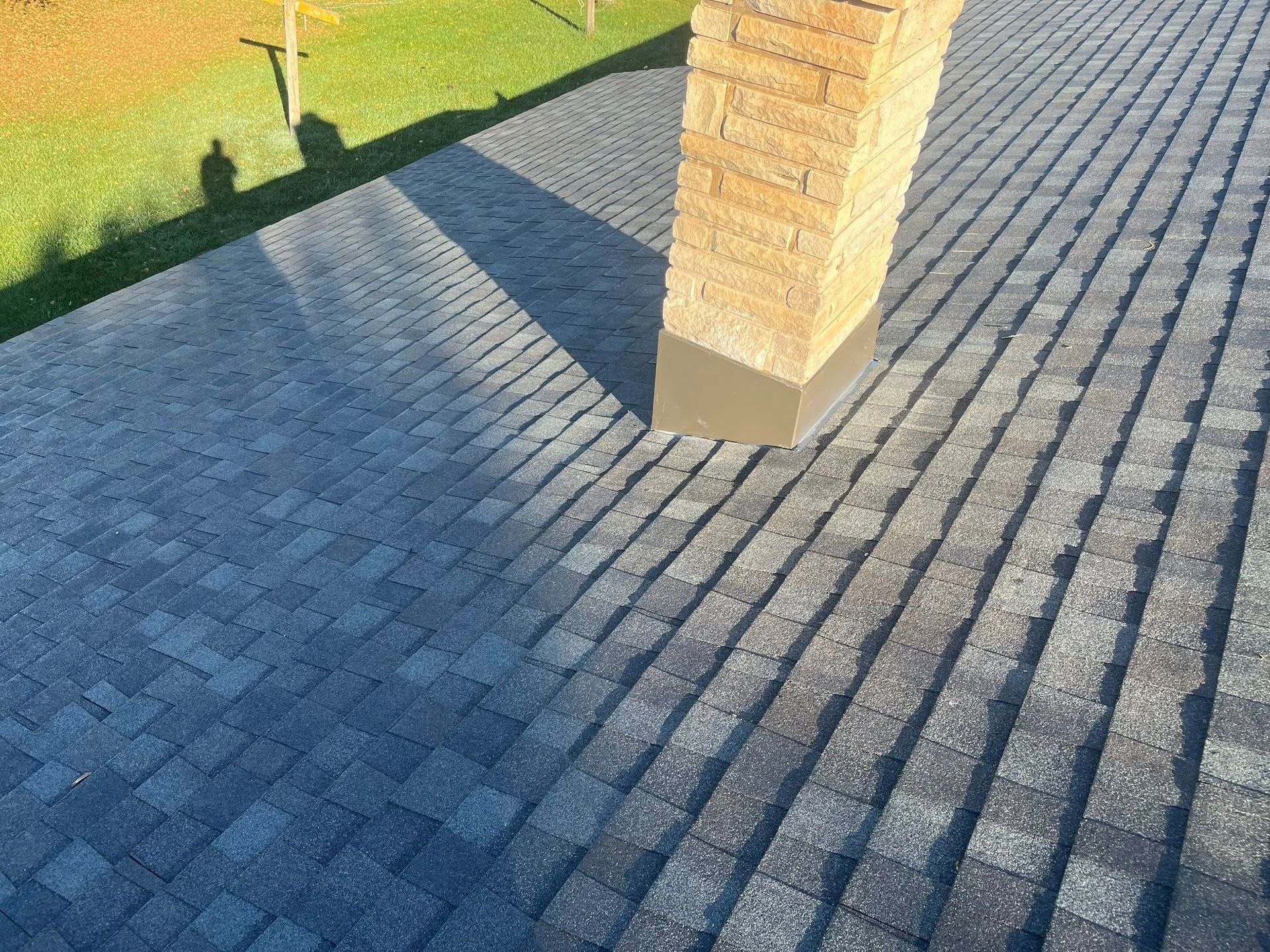 A tan brick chimney rises from a dark, gray shingled roof, casting a long shadow next to a patch of green grass.