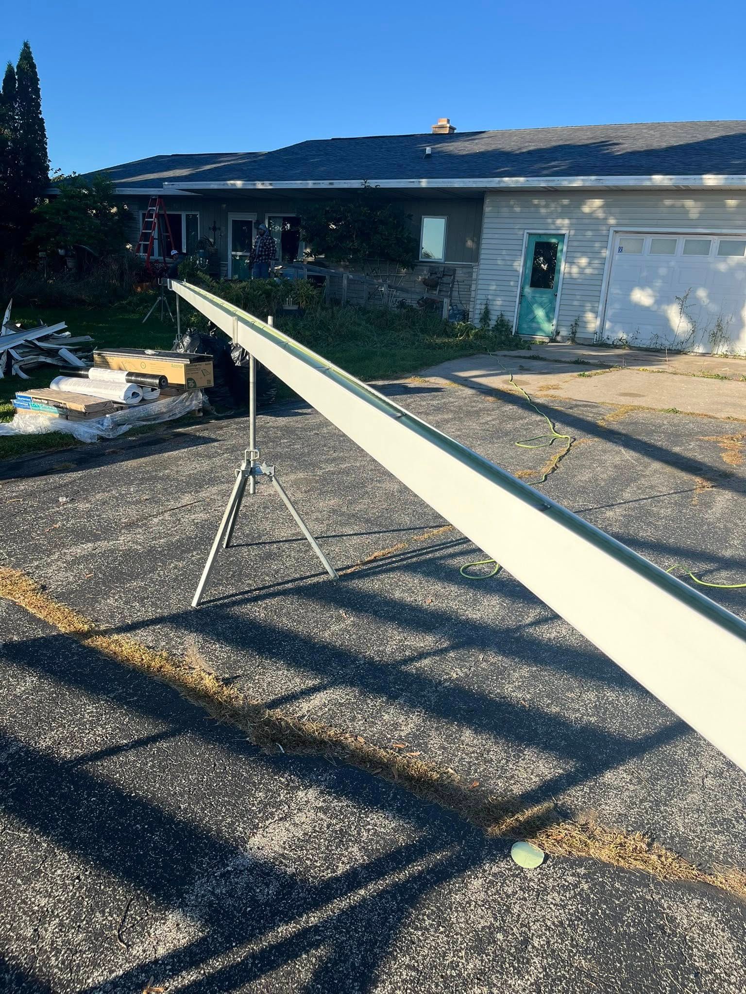 A long, light-colored gutter section rests on a tripod stand in a driveway in front of a house.