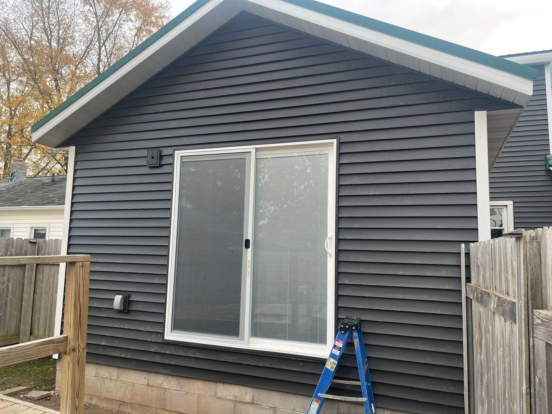 Exterior view of a gable-roofed home addition with dark gray horizontal siding, a sliding glass door, and a blue stepladder.