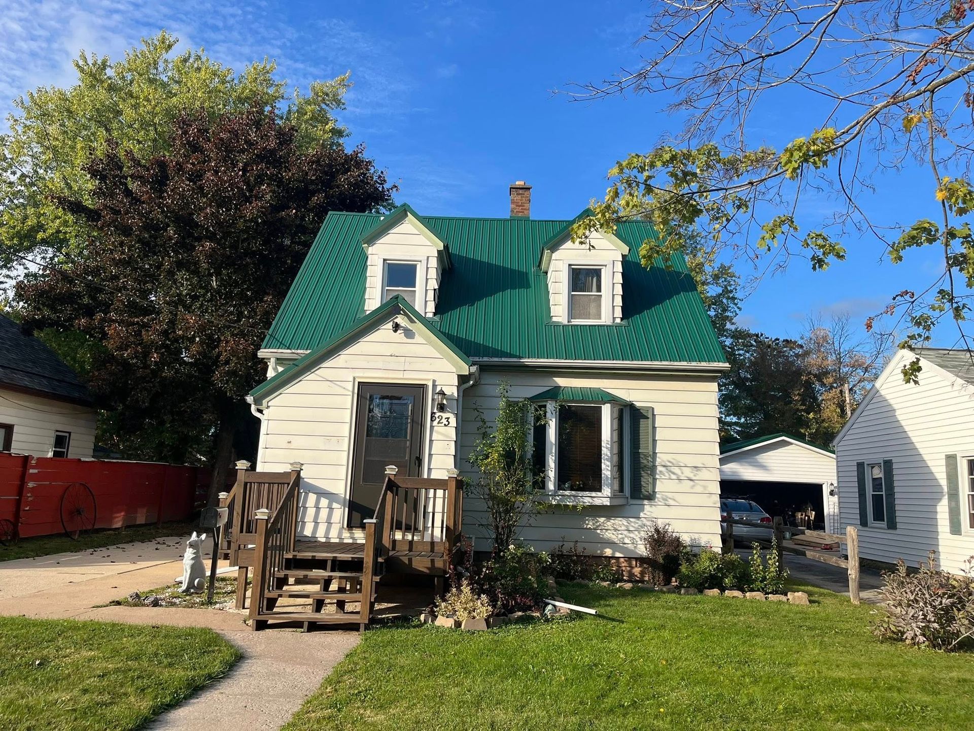 A two-story house with white siding and a green metal roof, featuring a front porch, dormer windows, and a grassy yard.