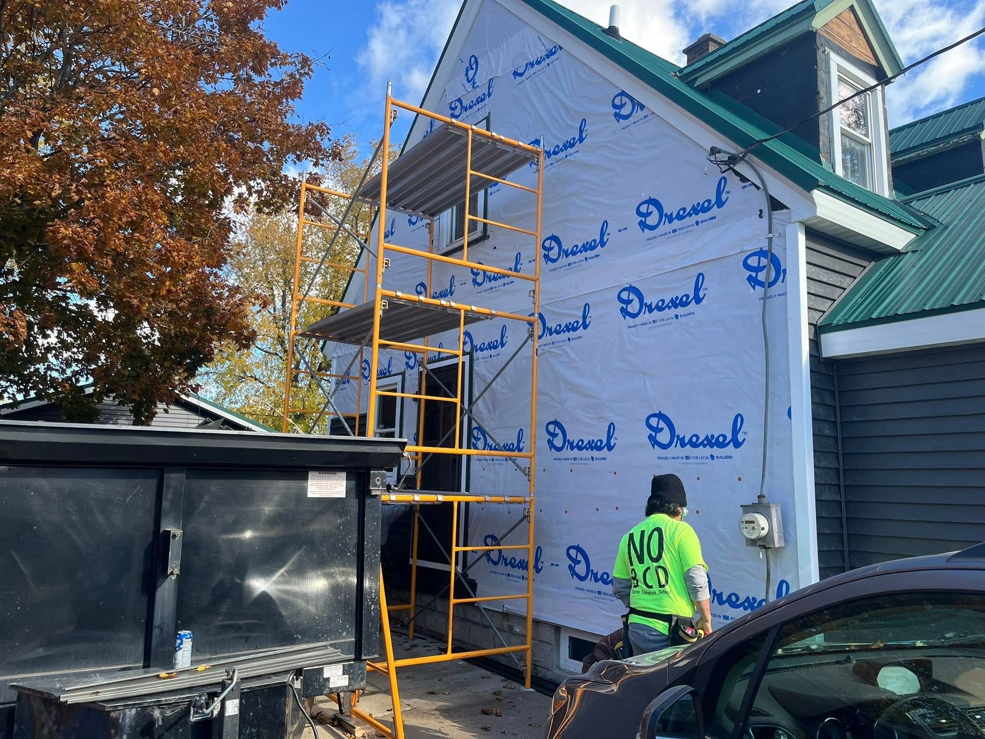 A worker in a bright green shirt stands next to scaffolding against the side of a house wrapped in blue house wrap.