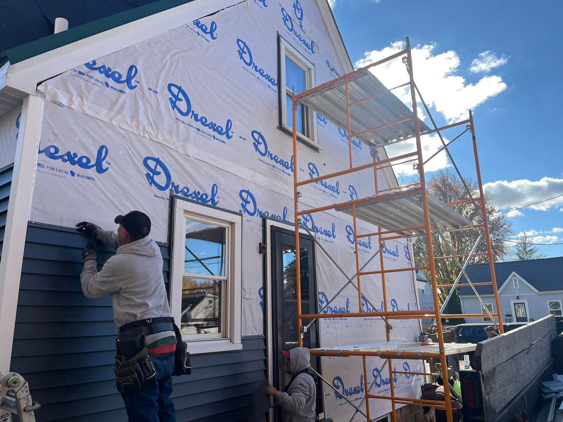 Two workers install dark gray siding on the exterior of a house under construction, using scaffolding on a sunny day.