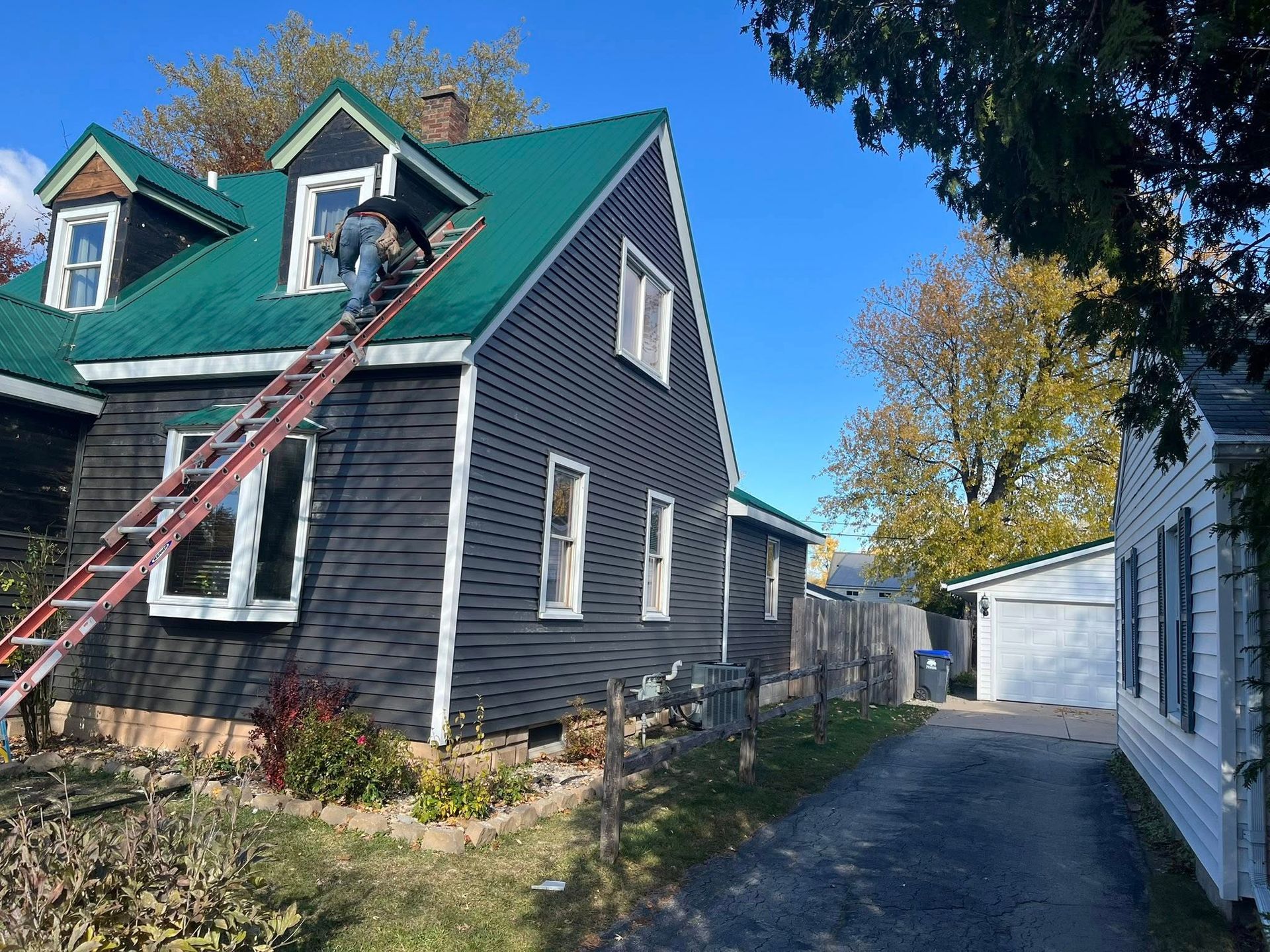 A worker on a ladder performs repairs on the green metal roof of a dark-sided house under a clear blue sky.