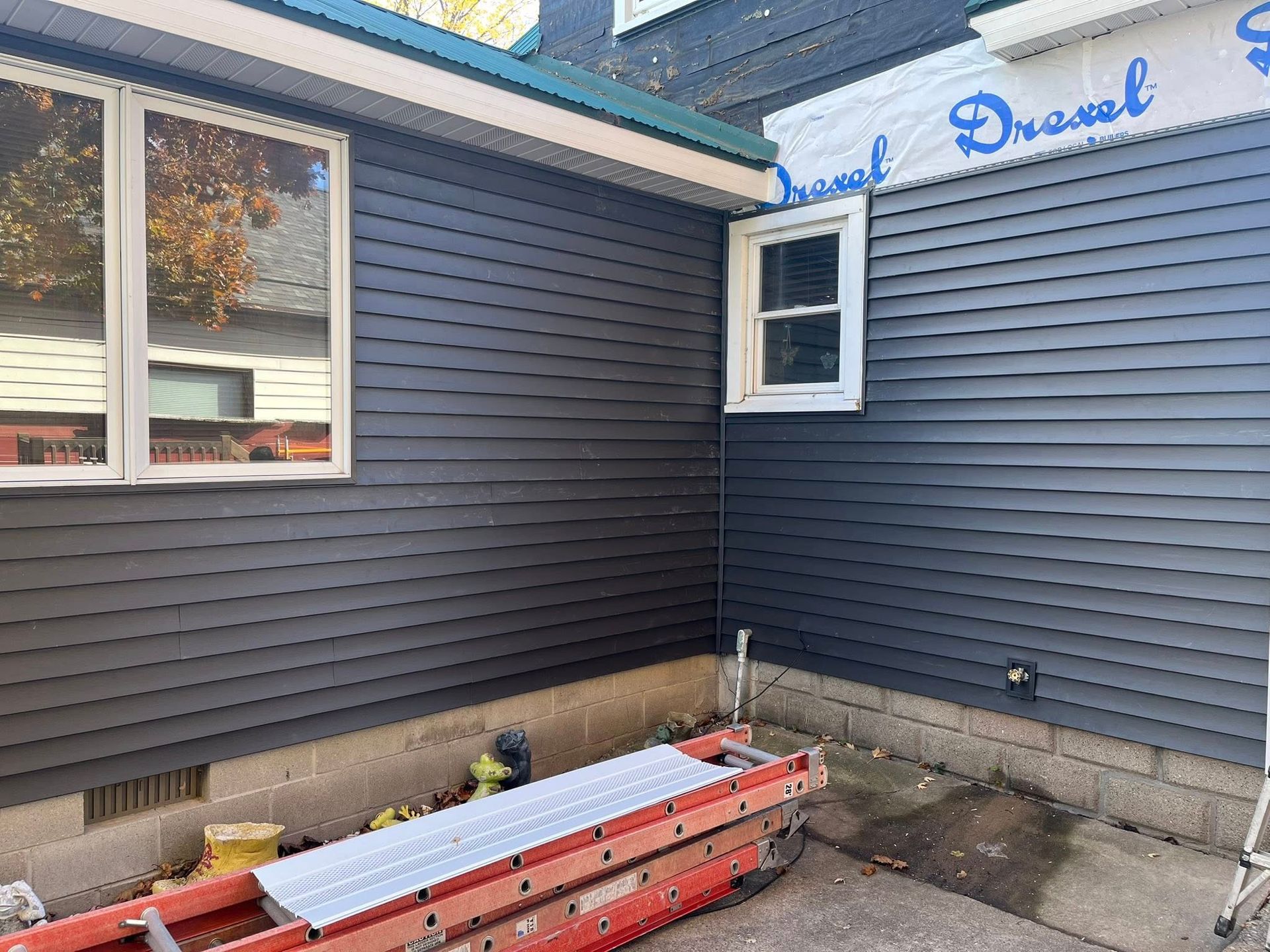 Exterior of a house under renovation with dark grey vinyl siding, new windows, and an orange ladder on the ground.