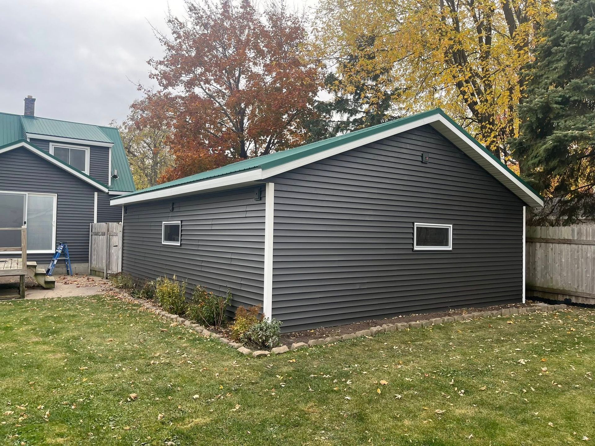 A detached garage with dark grey horizontal siding and a green roof, positioned in a yard next to a matching house.
