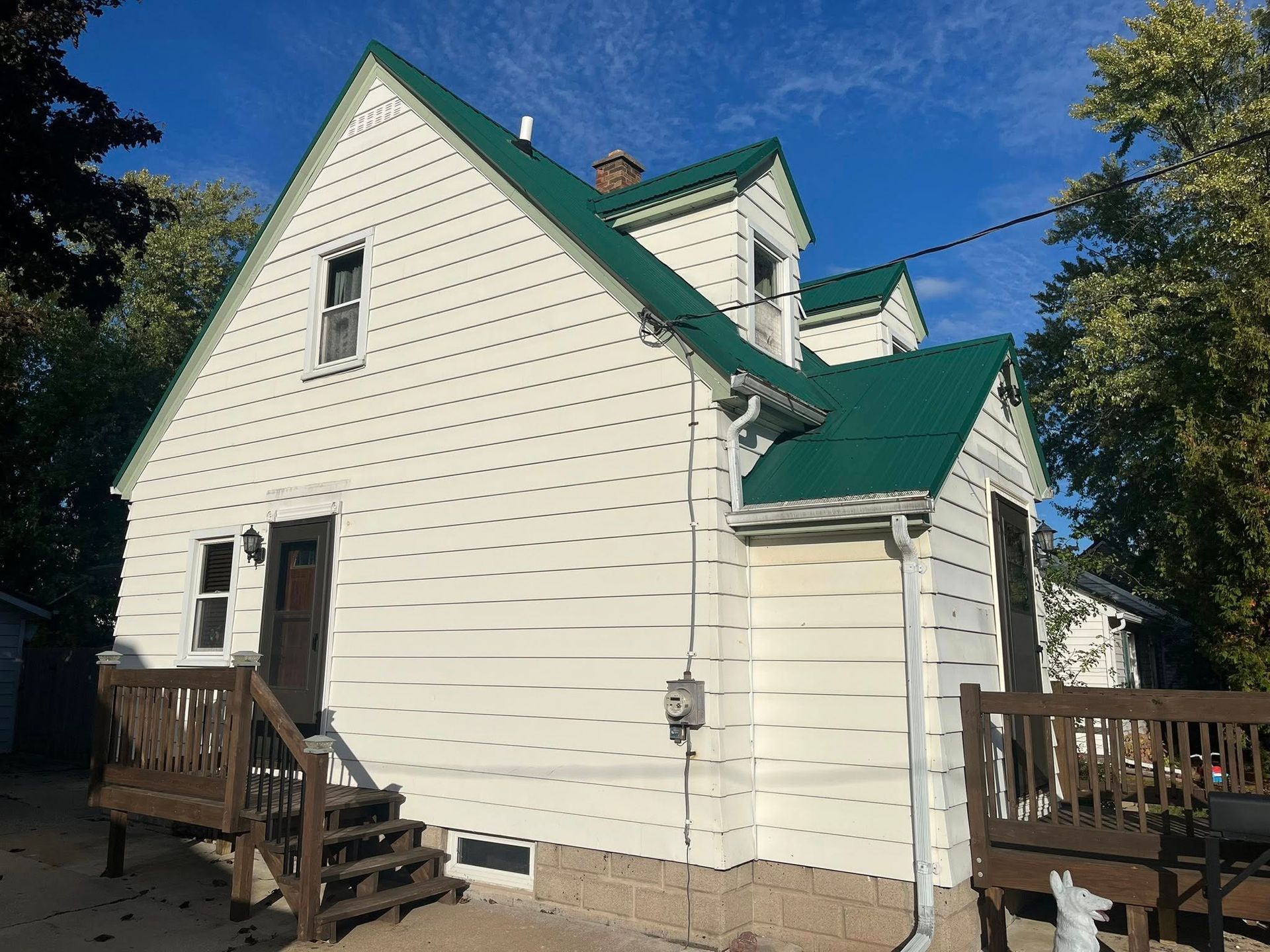 A white, two-story house with a green metal roof, a wooden deck, and wooden stairs against a blue sky.
