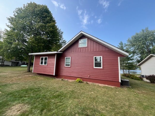 A side view of a red-sided house with white trim, set on a green lawn against a blue sky with a large tree.