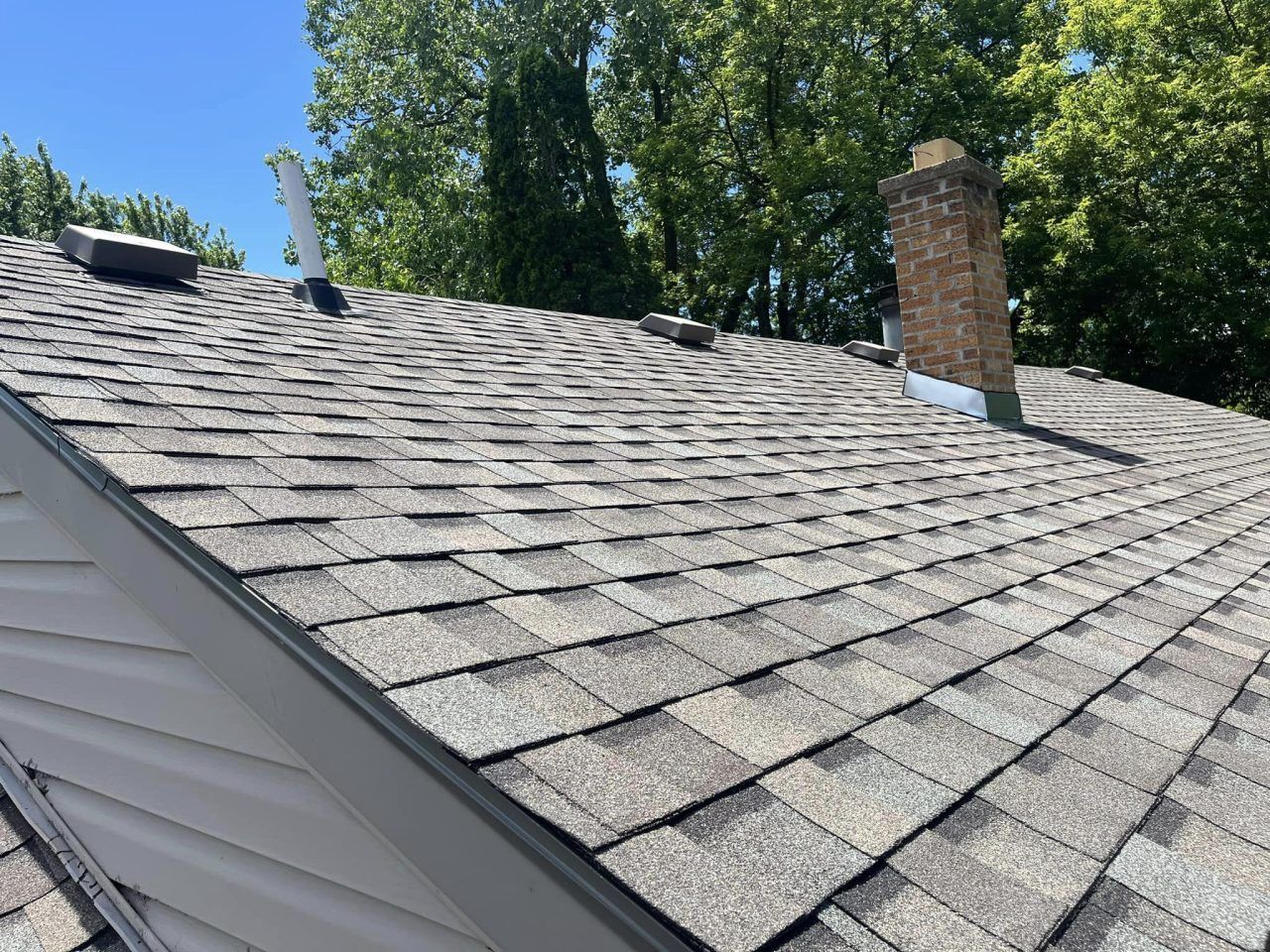 A roof with gray asphalt shingles, a brick chimney, and a white plumbing vent pipe under a clear blue sky.