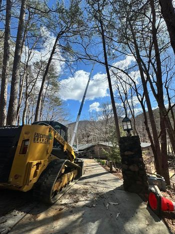 Yellow skid steer, crane reaching up, trees, blue sky, driveway, house.