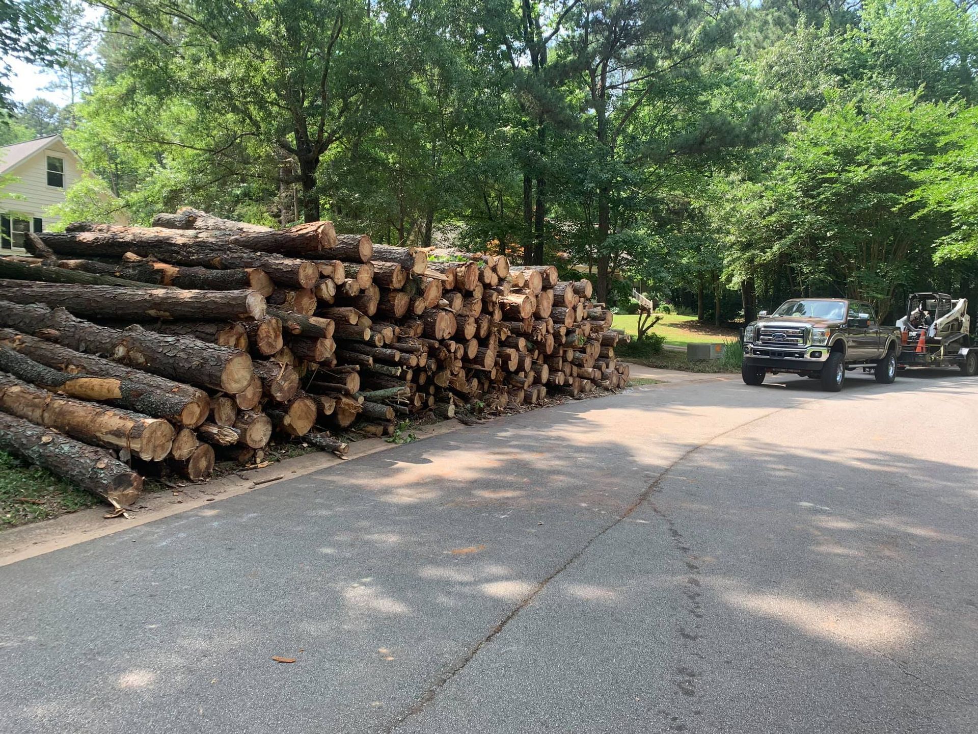 Pile of logs beside a road, truck with trailer in the background, trees and house visible, sunny day.
