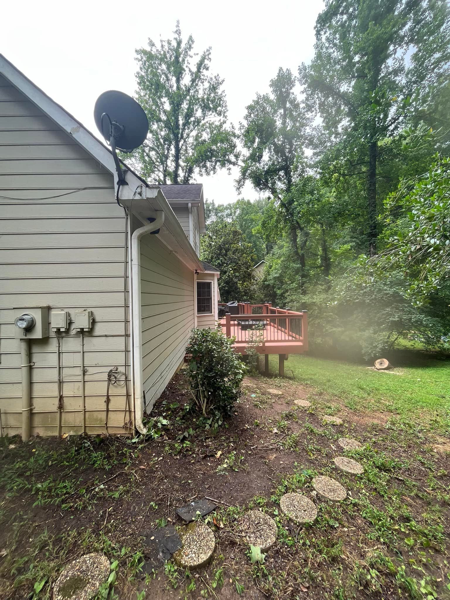 Side of a house with a deck. Beige siding, satellite dish, and green foliage. Wooden deck with red stain.