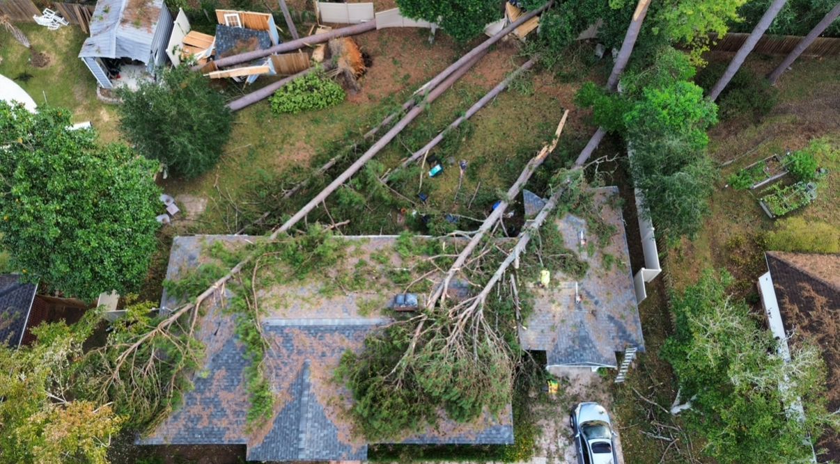 Aerial view of a house with several large trees fallen on top, possibly after a storm. Debris scattered in yard.