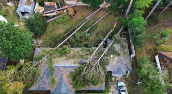 Aerial view of a house with several large trees fallen on top, possibly after a storm. Debris scattered in yard.