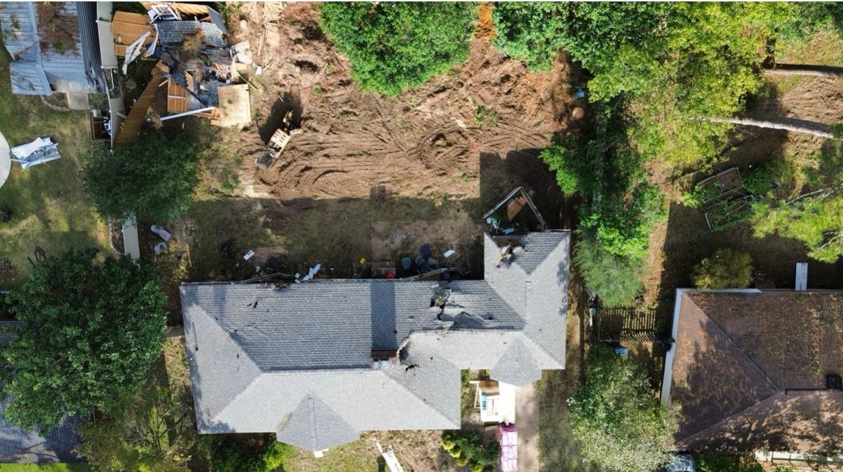 Overhead view of a house with a damaged roof, debris, and a cleared area for construction. Trees surround the property.
